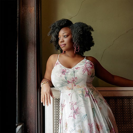 Alicia Olatuja stands staring out the window in a white floral dress.