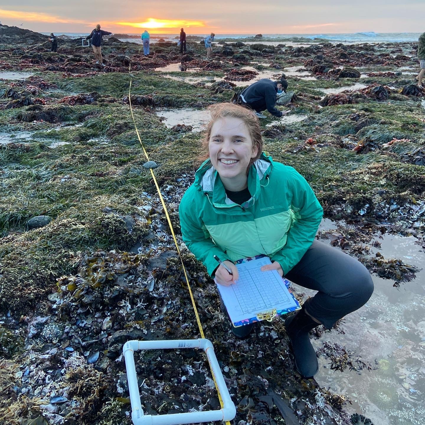 Student Audrey Sarin smiles for a photo while kneeling down and holding a clipboard at the beach. 