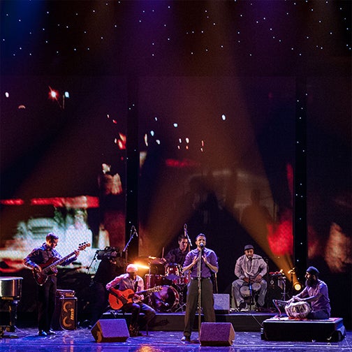 Aditya Prakash is pictured on stage with blue and red lights behind him as his band stands behind him.