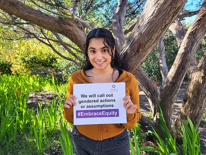 A female student stands in front of a tree holding a piece of paper reading "We will call out gendered actions or assumptions #Embrace Equity"