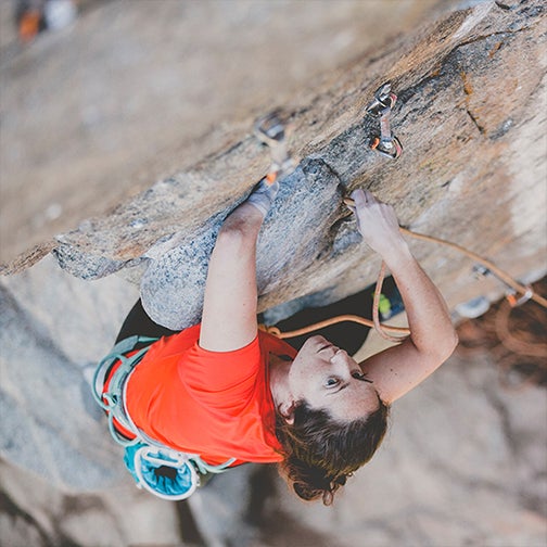 This picture is shot from a rock above Maureen Beck as she climbs up a rock with her arms reaching for the camera. She is tied in with ropes and wearing a bright orange-red shirt. 