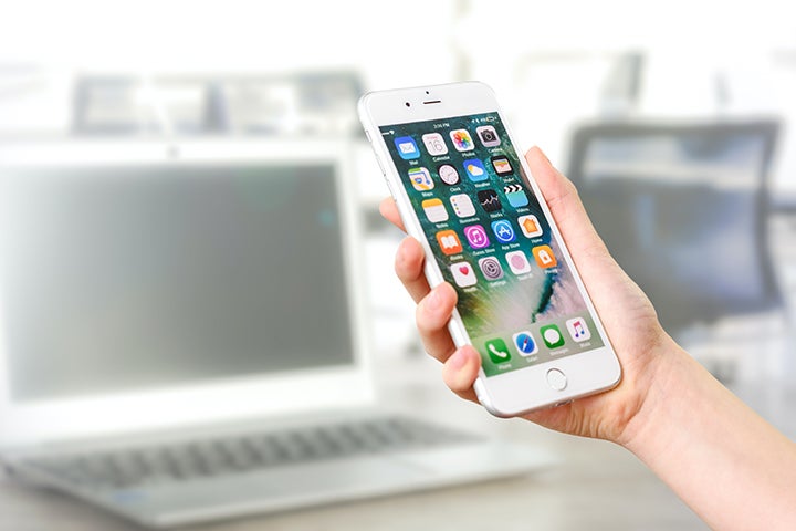 A hand holds an iPhone while a laptop sits on a desk in the background.