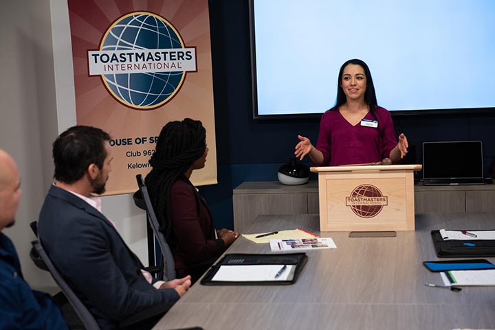 An individual stands behind a podium and speaks to a group of people who are seated around a table, with a Toastmaster International sign displayed behind them.