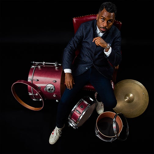  Shot from above, the picture shows Nate looking up at the camera in a suit, sitting in a red chair surrounded by his drum set. 