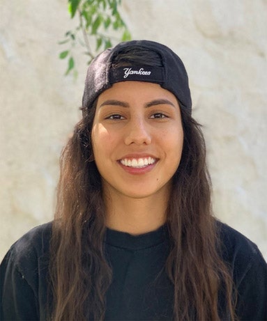 A young woman in a T-shit and black baseball cap smiles into the camera