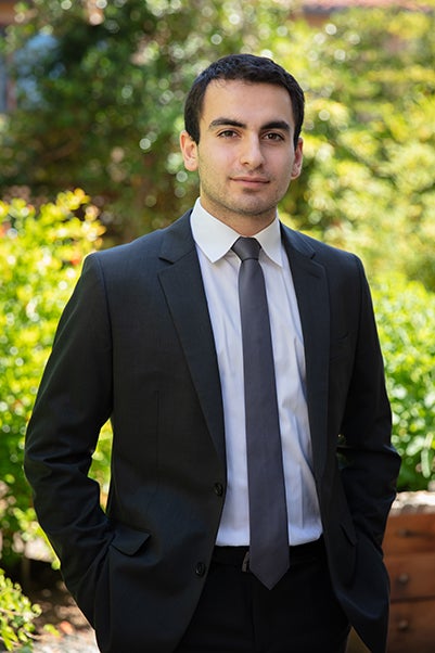 A man in a black suit and tie poses with both hands in his pockets. 