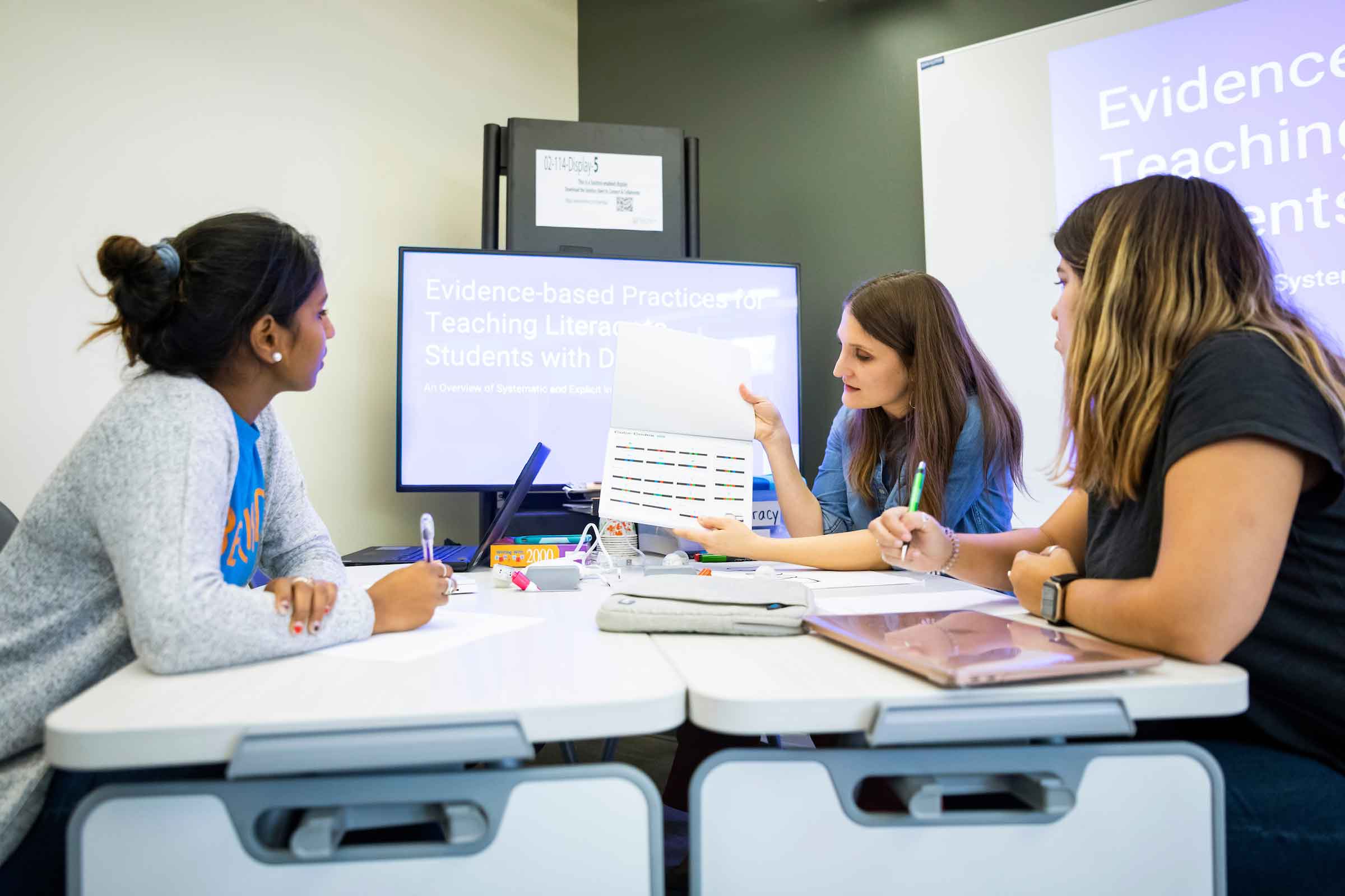 Three women gather around a screen and are talking.