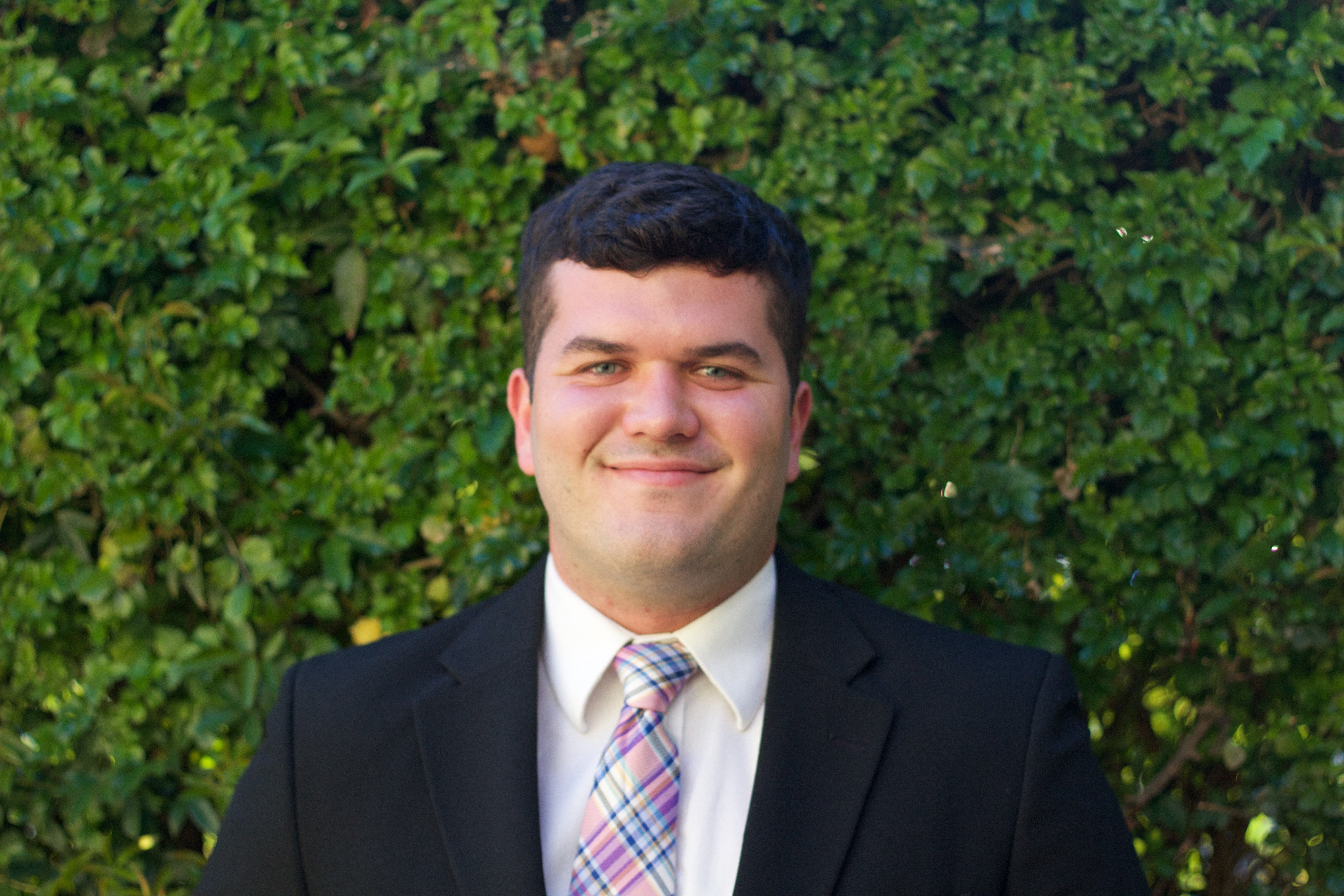 A man in a white button down, pink plaid tie and black suit jacket smiles in front of a shrubbery.