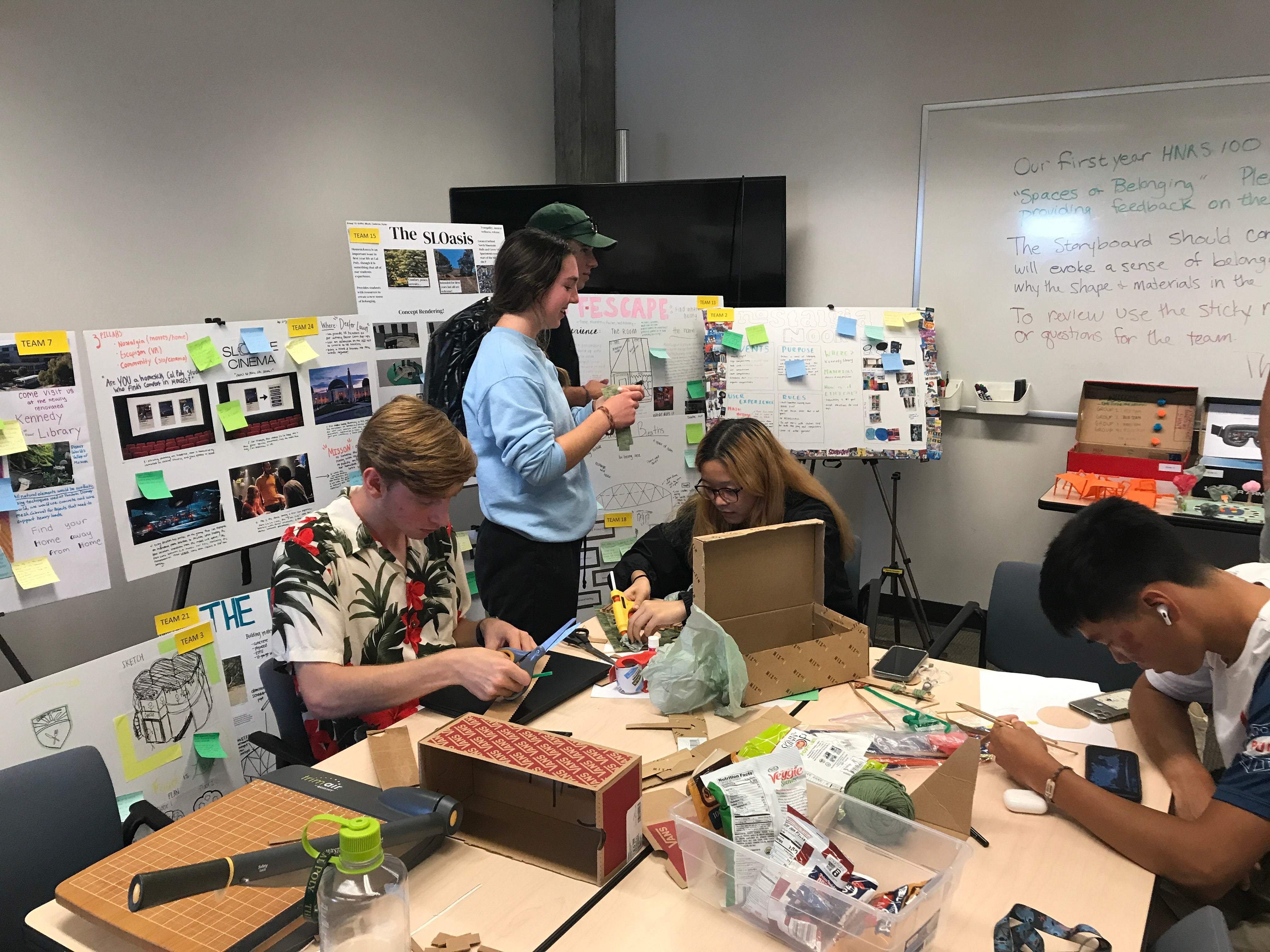 Students gather around a table covered in poster boards and sticky notes as they design a space of belonging.