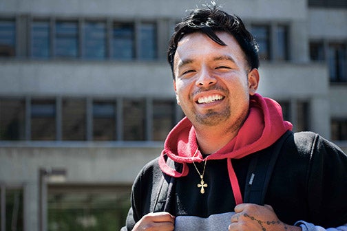 A student in a sweatshirt and backpack smiles in front of the Kennedy Library