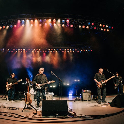 A stage with colorful lights from above shines down on three guitarists as they sing together.