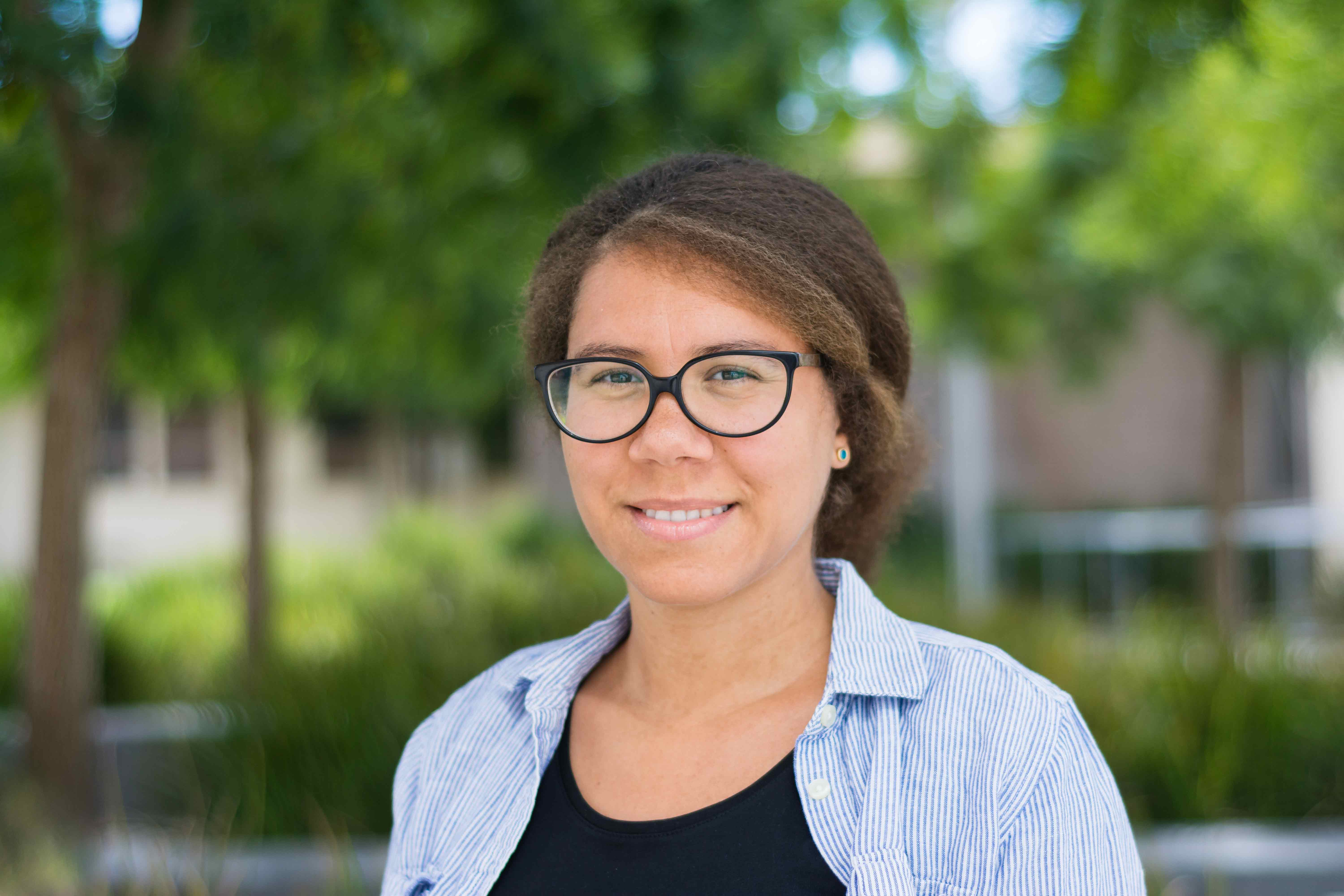 A woman with black frame glasses and light brown hair pulled up smiles and wears a blue and white striped button down over a black shirt.