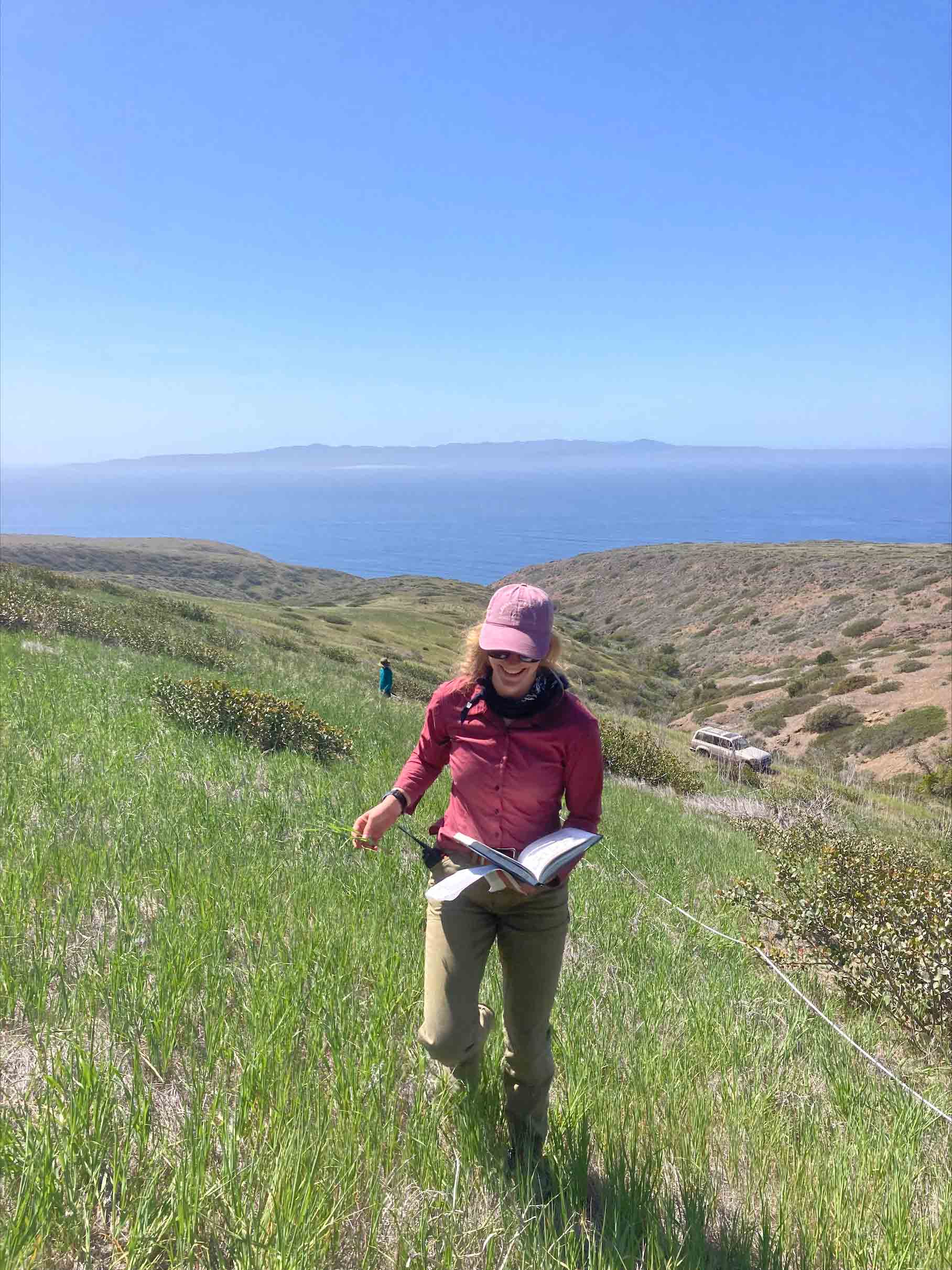 A woman in long pants and a jacket smiles while she holds a large notebook and walks up a grassy hill. 