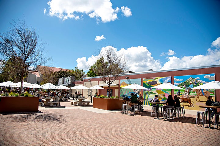 Campus community members sit at tables on Via Carta Road near Campus Market, with a colorful mural on the wall in the background.