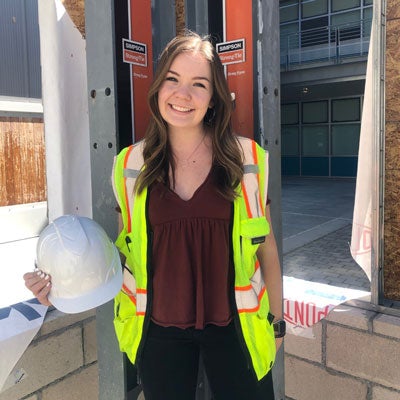 A woman in a reflective vest stands with a hard hat in front of a construction site.