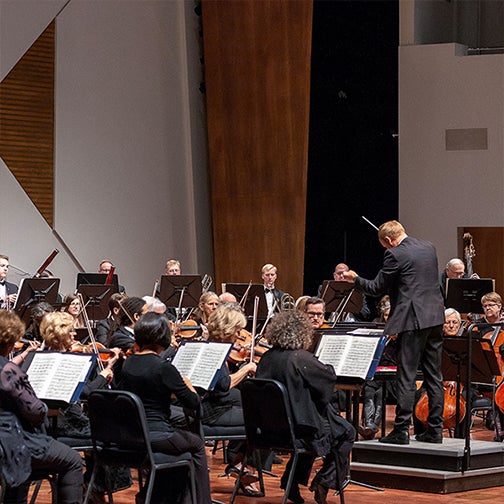 A picture shows the stage with the conductor’s arm raised and the symphony around him, their music sheets visible as they sit in seats playing their instruments.