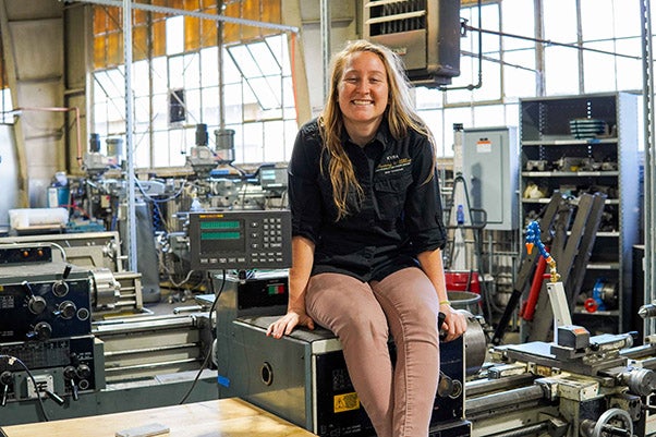 A young woman smiles while sitting on a tool bench in a workshop