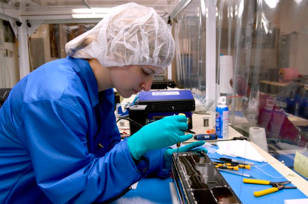 A woman in a blue coat, teal latex gloves and a white scrub cap uses a screwdriver to work on the ExoCube 2 CubeSat.