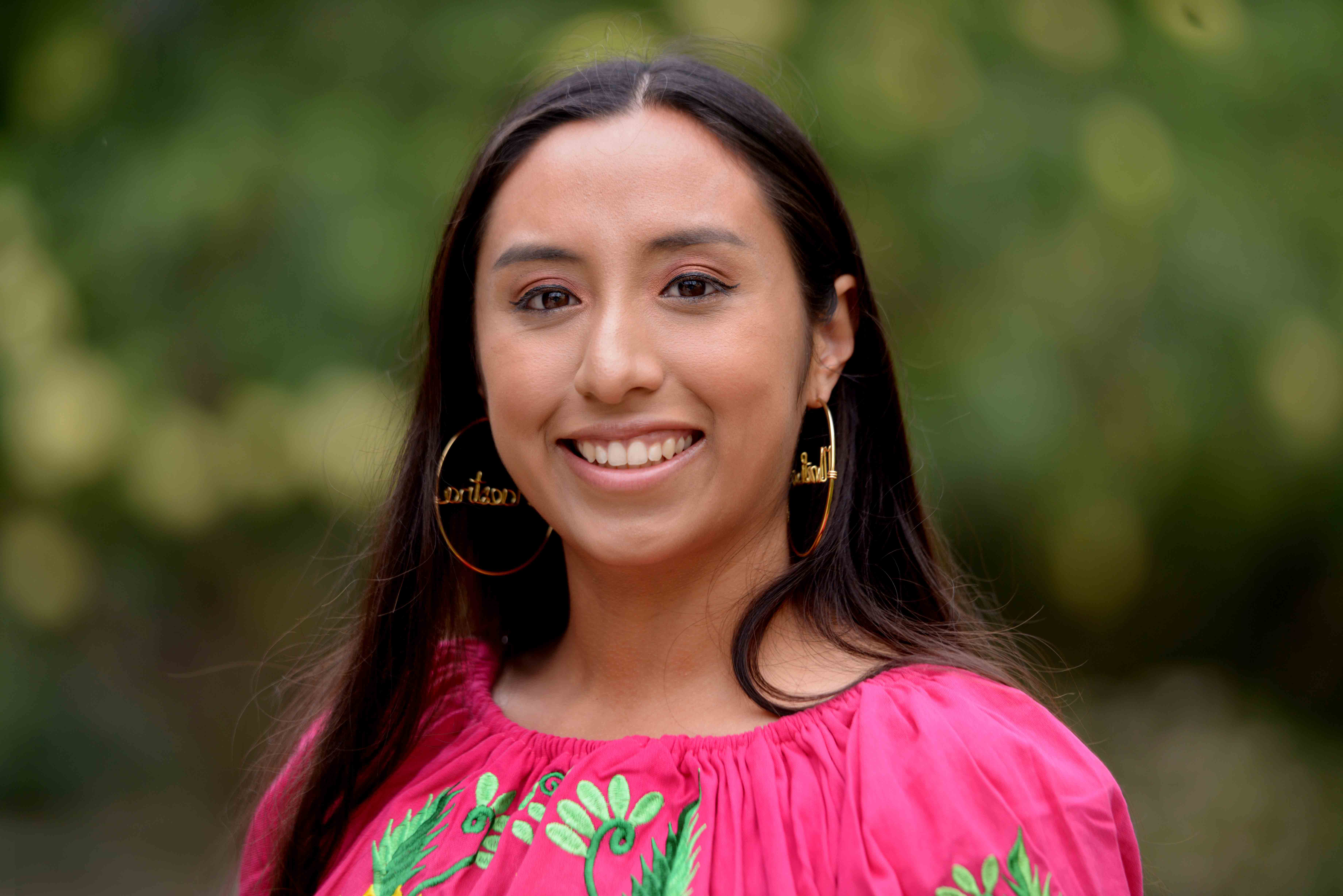 Gloria Sevilla wears an embroidered pink blouse and gold hoop earrings as she smiles for her headshot.