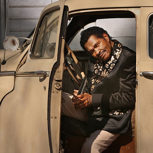 Singer in a black suit with floral accents sits in an old tan truck with the door open holding an instrument.