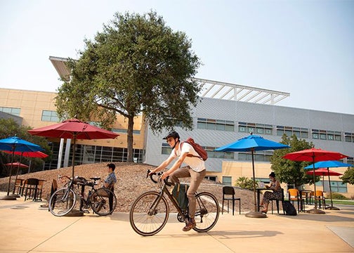 A male student rides his bike past some campus engineering buildings