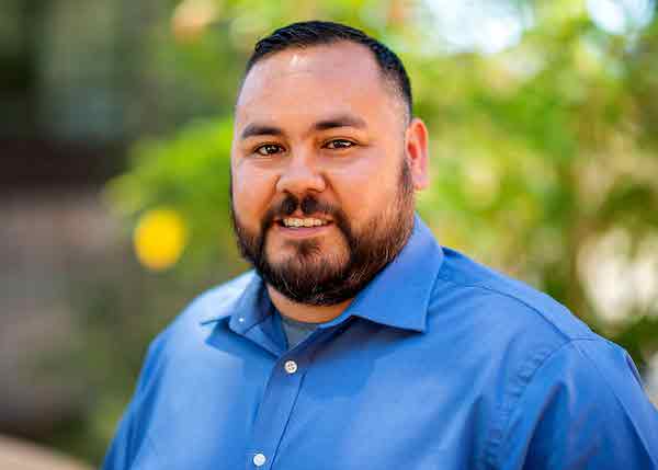 A man with short black hair and a black beard smiles at the camera and wears a blue long sleeve button down.