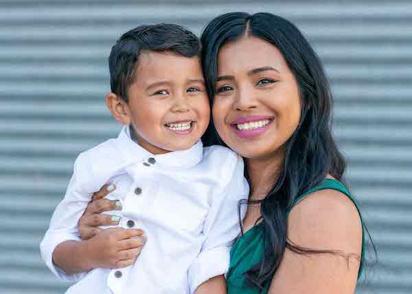 A black-haired woman wearing pink lipstick and a green sleeveless top smiles and holds a little boy wearing a white long-sleeve button down