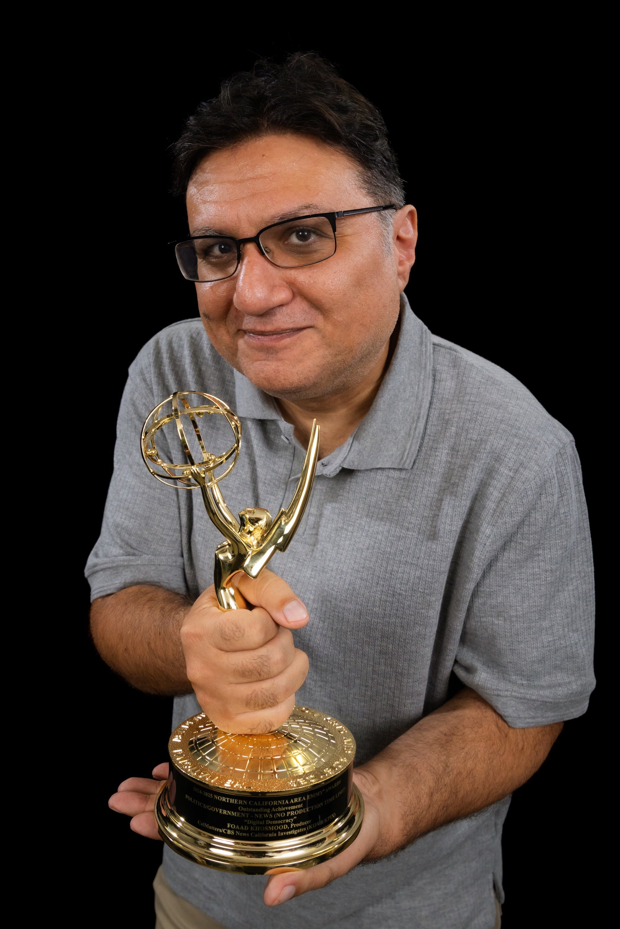 A professor in a gray shirt holds an Emmy statuette.