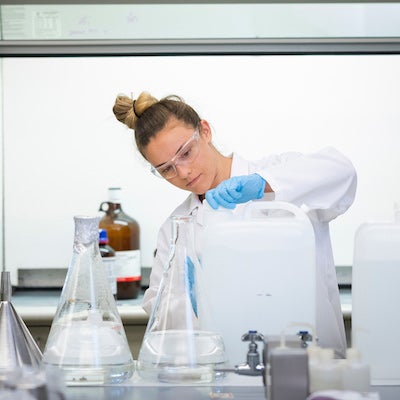 A student in a white lab coat pours liquid into a class container