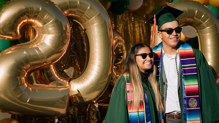 Two students in green graduation robes smile in front of gold balloons that say '2020'