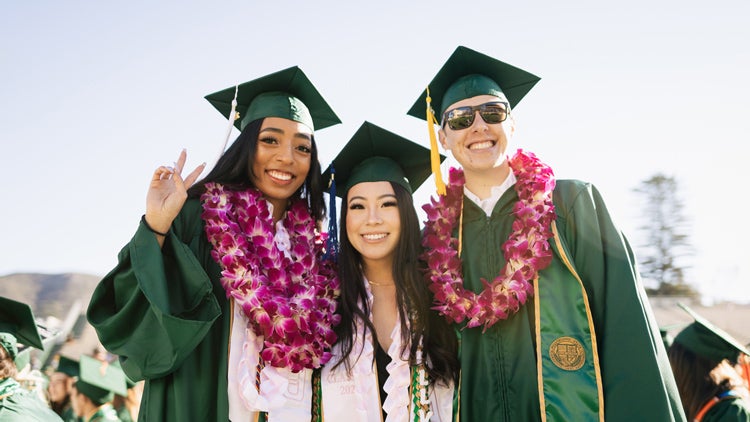 Three students in graduation robes and hats embrace and smile at a commencement ceremony