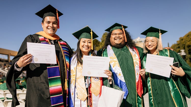 Four students in cap and gown hold commencement certificates and smile