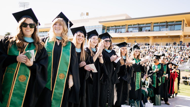 A row of graduates wearing black robes smiles before a stadium filled with supporters on a sunny day