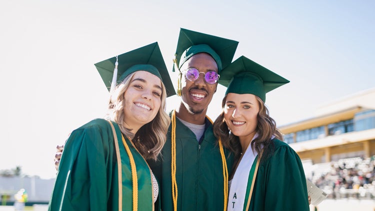 Three students in cap and gown embrace at a commencement ceremony in a stadium