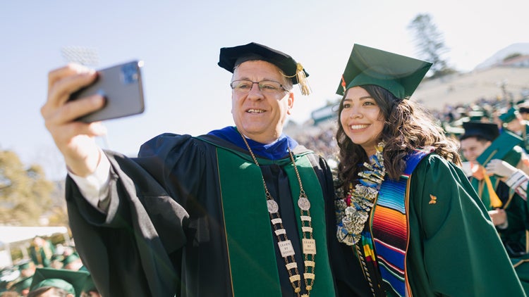 A faculty member and student take a selfie while wearing graduation robes