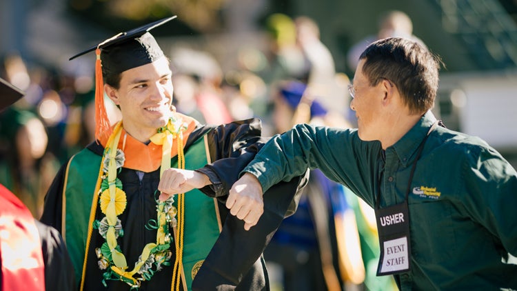 A graduate in cap and gown and a staff member bump elbows