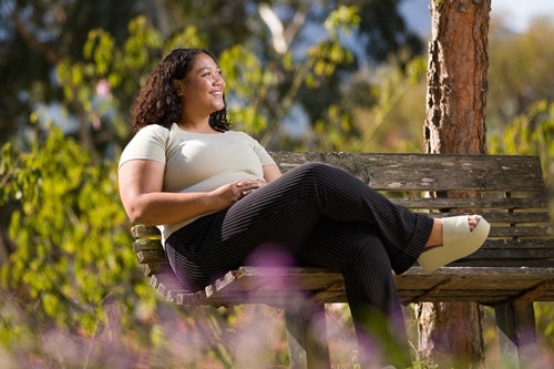 A person smiles while wearing a green shirt and black pants sitting on a bench under a tree 