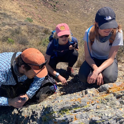 Three student researchers wearing hats kneel while examining lichens growing on a rock