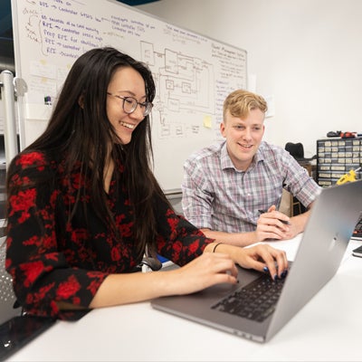 Two students sit at a computer with a white board covered in black writing behind them