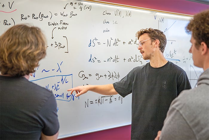 Two students and a professor look at math equations on a whiteboard.