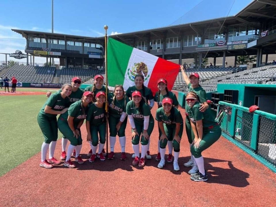 Female softball players from Team Mexico wear green uniforms and pose for a photo on the field, waving a Mexican flag behind them.