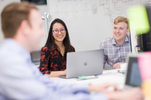 Three students smile and talk as they gather around their laptops at a table.