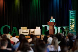 Dolores Huerta stands behind a lectern on stage at Chumash Auditorium. The banner next to her says ¡Adelante!, which is the name of the symposium she spoke at. 