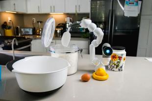 In a kitchen, a robotic arm with a glowing sensor eye prepares to scoop flour out of a jar into a bowl.