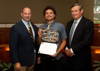 A young man in a shirt and tie holds a certificate while standing between two older men in black suits