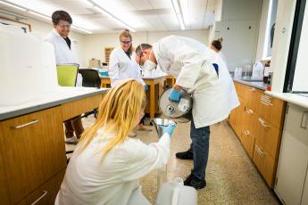 A man in a lab coat and mask pours hand sanitizer into a funnel held by a woman in a white lab coat. 