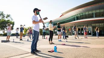 A dozen marching band members wearing face coverings play their instruments in front of the performing art center