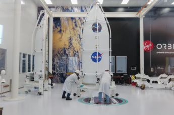 Two people in white coats and hard hats inspect a white payload vessel with NASA logo on the front inside the Virgin Orbit facility. 