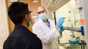 A professor wearing a mask demonstrates a chemistry experiment to a student in a lab