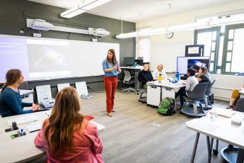 A woman in red pants and a blue top stands in the middle of a classroom surrounded by students.